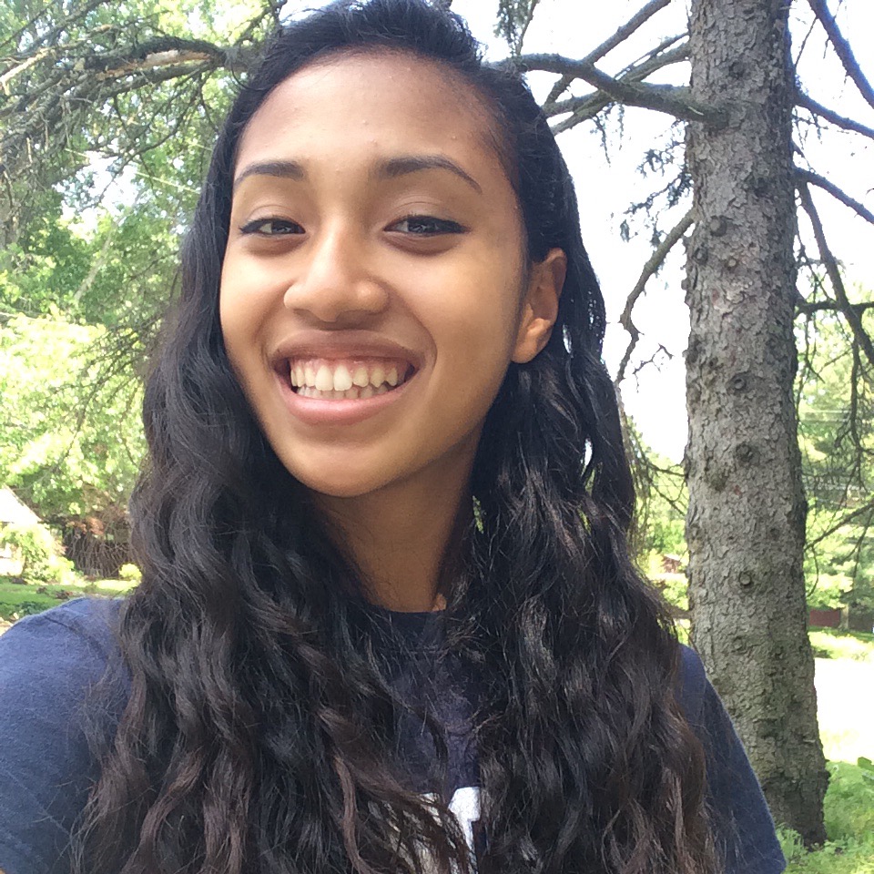 A tan, pretty girl with dark brown curly hair. She is smiling.