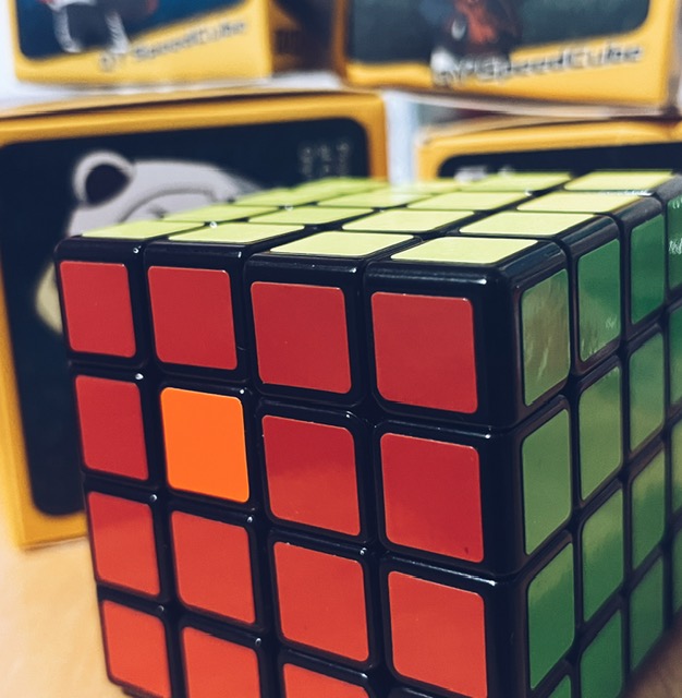 Image of a 4 by 4 rubiks cube with the red and green sides facing the camera and the yellow one on the top. There is one orange center square at the top left where a red one should be.